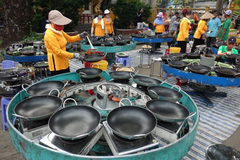Outdoor market scene with vendors in vibrant attire preparing food at multiple cooking stations.
